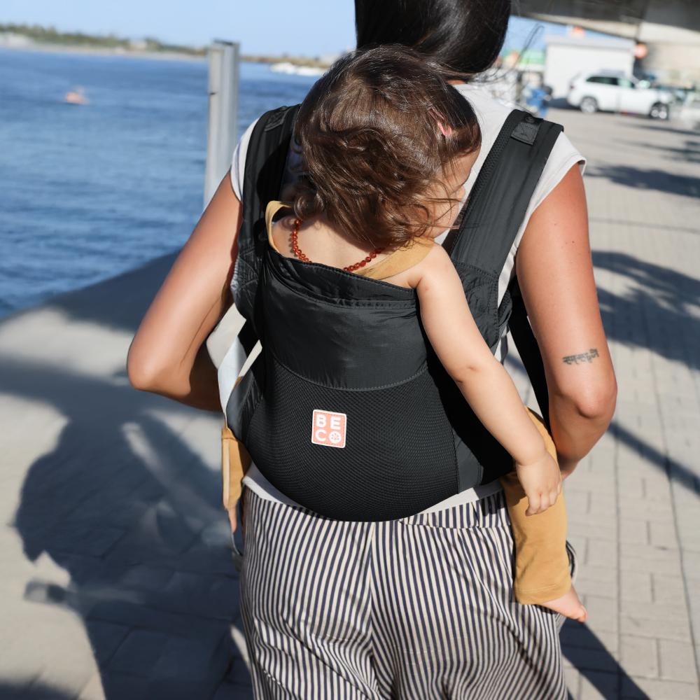 Person carrying a toddler in a black light mesh baby carrier by a waterfront.