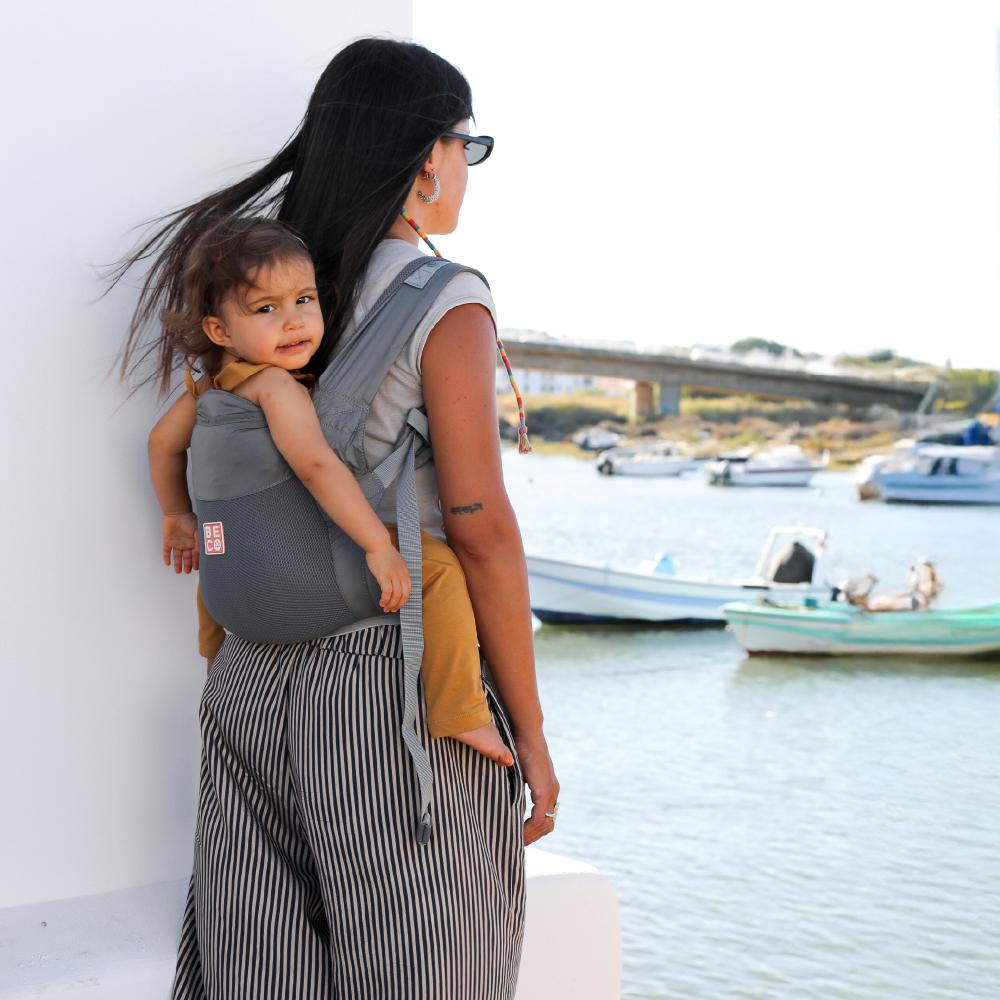 Woman carrying a toddler on her back in a gray mesh summer baby carrier by a waterfront with boats in the background.