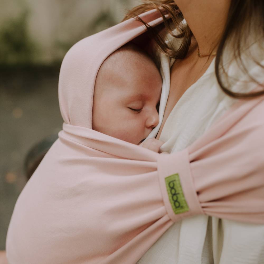 Newborn baby wrapped in a pink hybrid wrap carrier with a visible brand logo, held by a person outdoors.