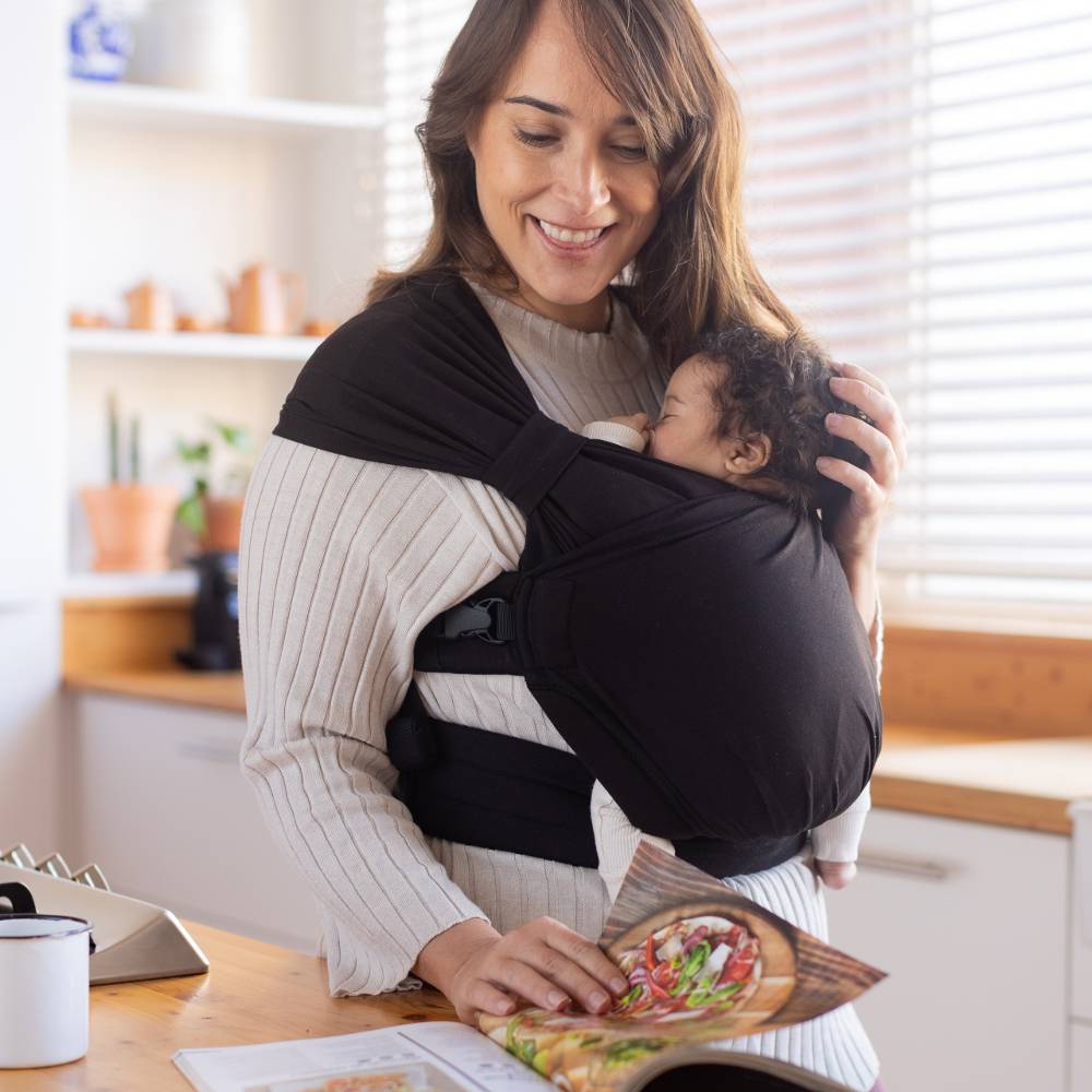 Woman holding a baby in a Boba Bliss Carrier while reading a magazine in a kitchen.