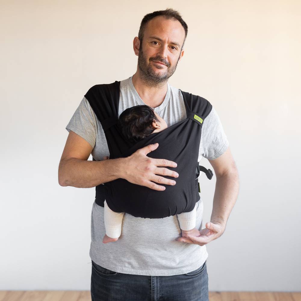 Man holding a baby in a black baby carrier against a plain background