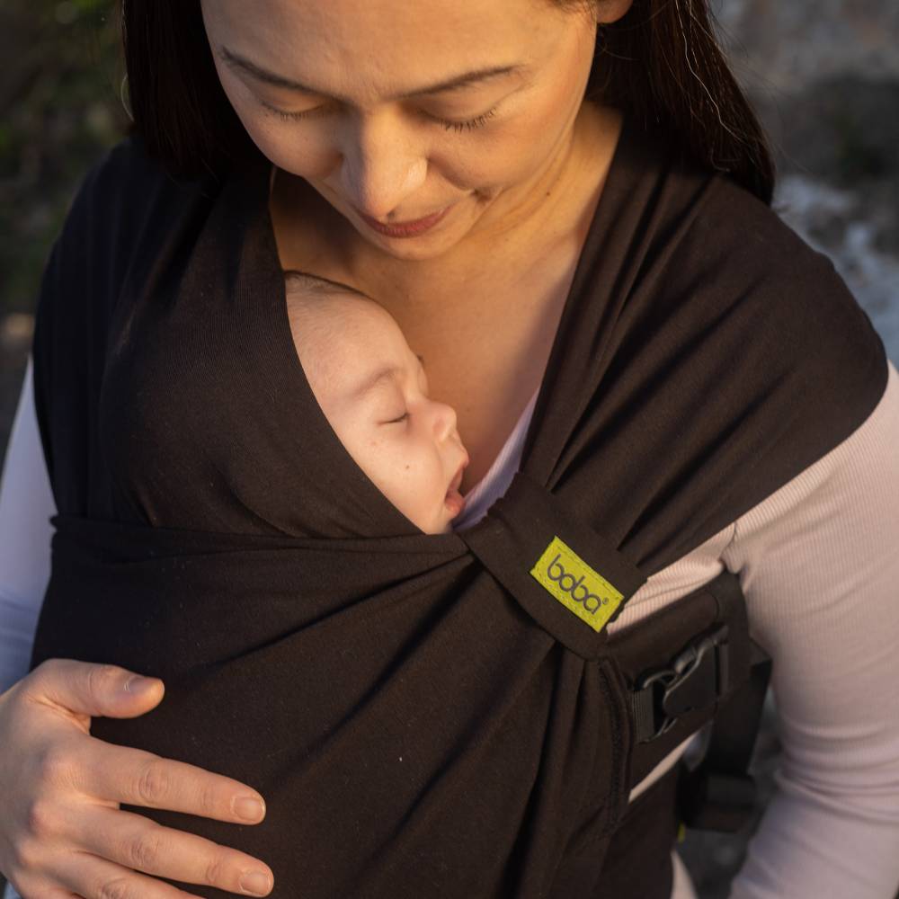 Woman holding a baby in a brown baby carrier with 'Boba' branding outdoors.