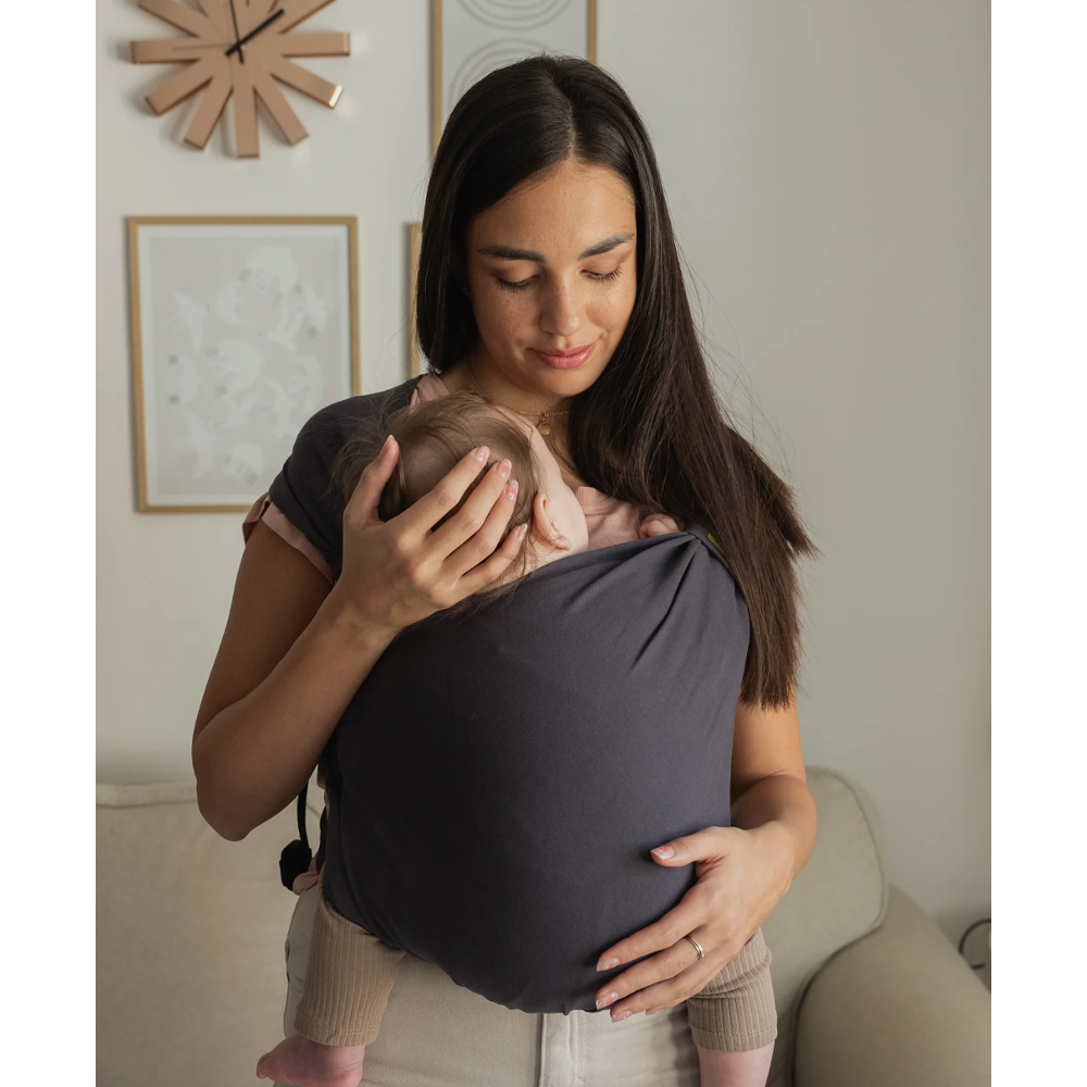Woman holding a baby in a dark gray sling in a home setting.