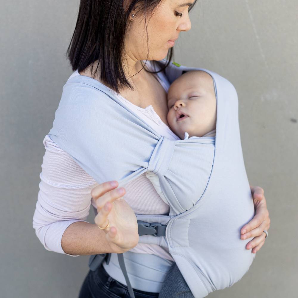 Woman holding a baby in a light purple baby carrier against a plain background