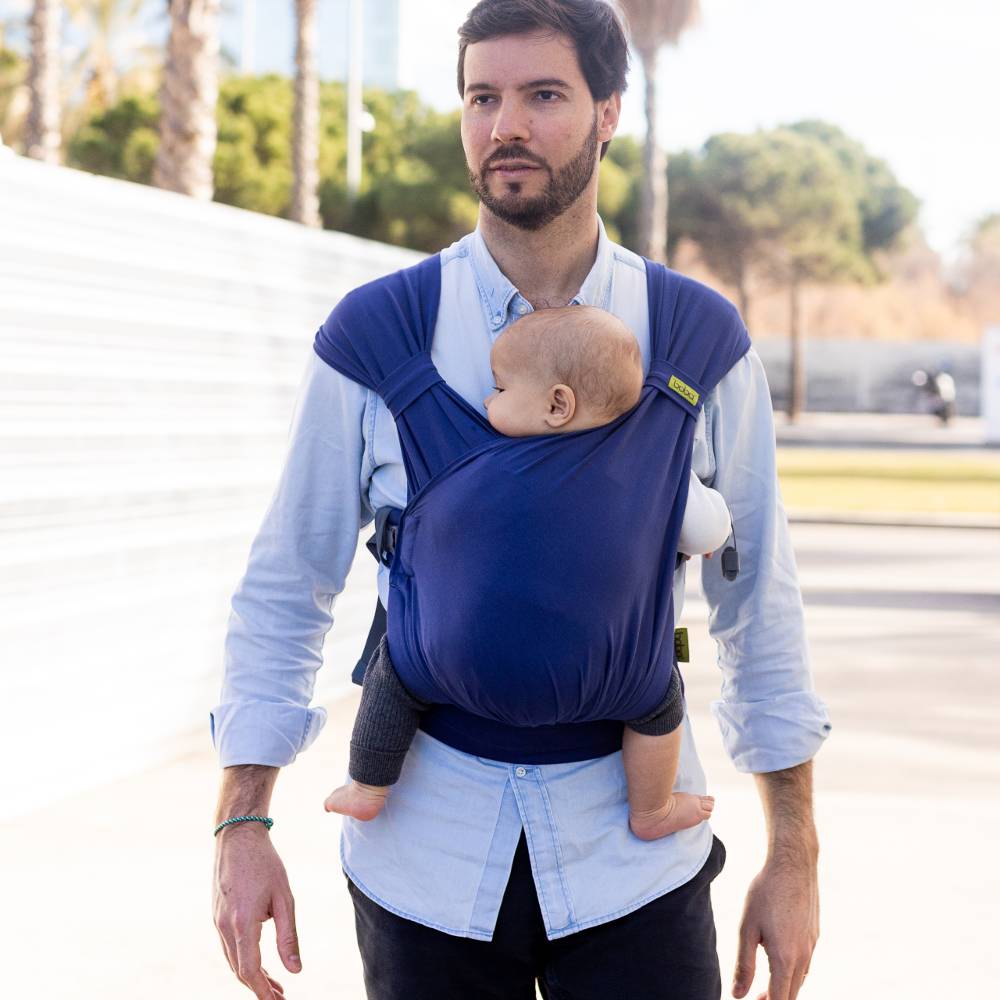 Man carrying a baby in a blue sling outdoors on a sunny day.