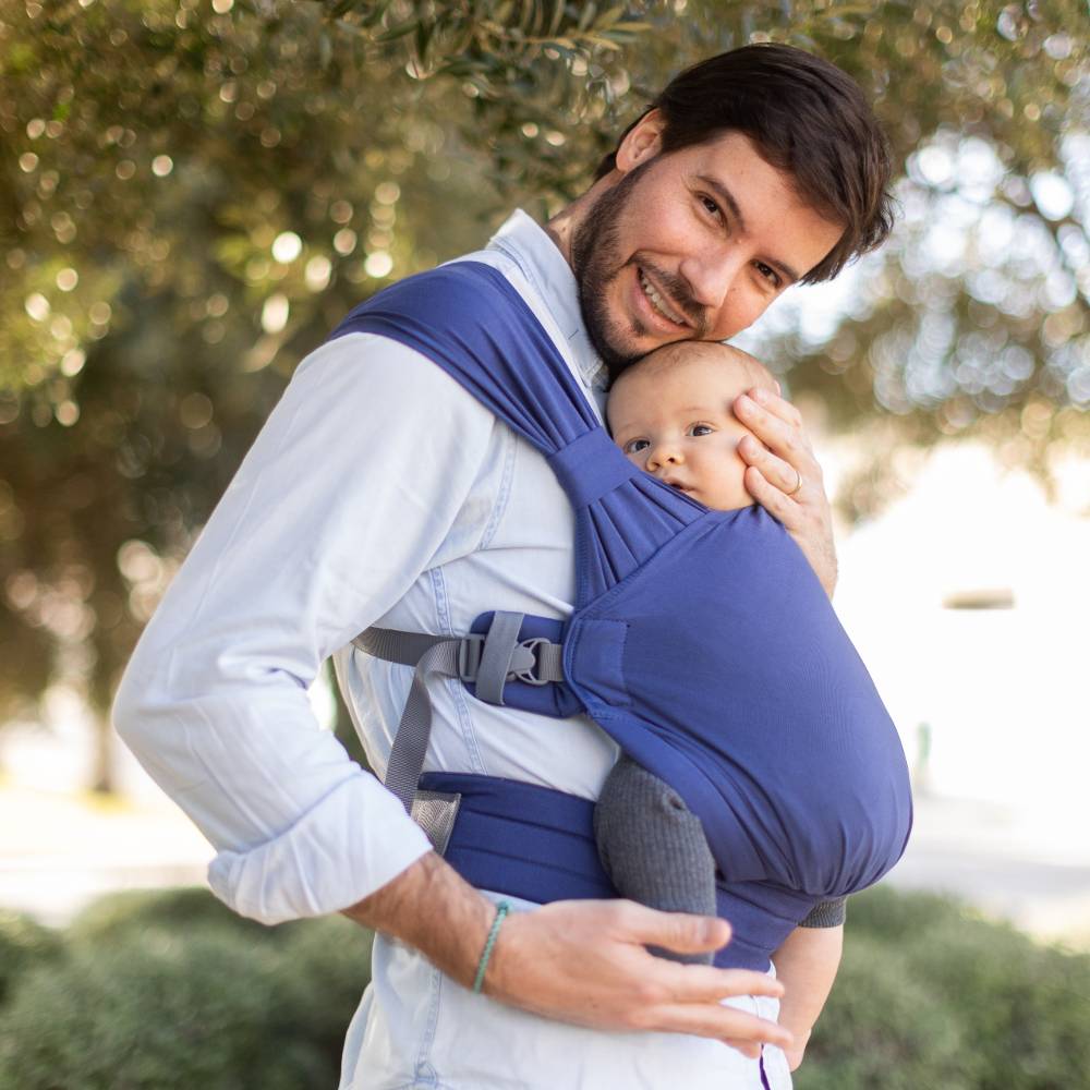 Man carrying a baby in a blue sling outdoors with greenery in the background