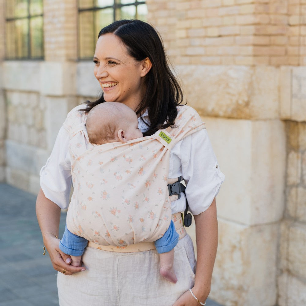 Woman carrying a baby in afloral Boba Bliss hybrid wrap carrier against a stone wall.