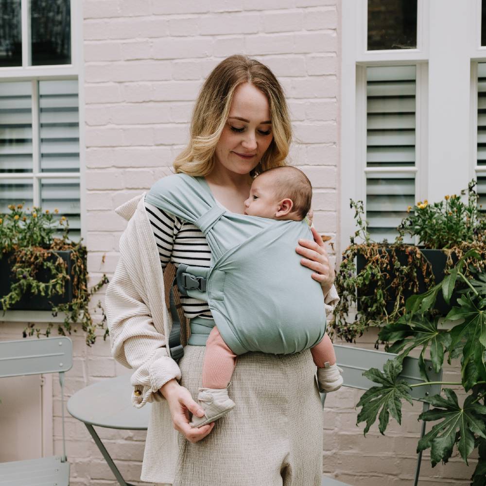 Woman holding a baby in a hybrid buckle carrier sling against a light-colored wall with plants.
