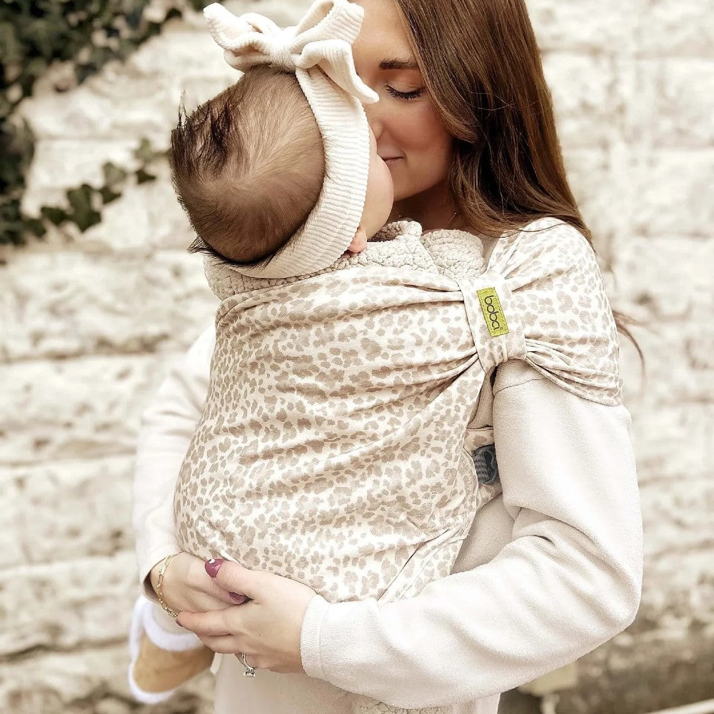 Woman holding a baby in a leopard print baby carrier against a blurred natural background