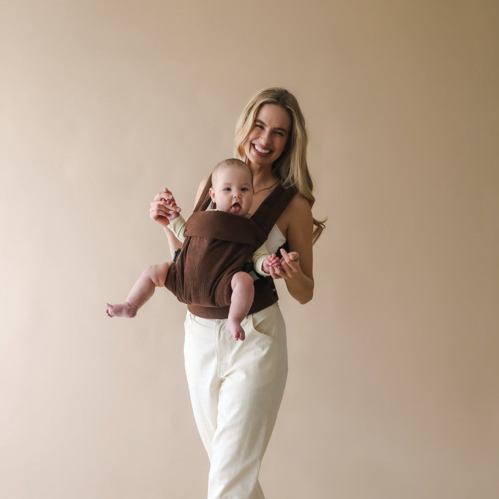 Woman holding a baby in a brown Boba Auri carrier in the front facing position against a beige background