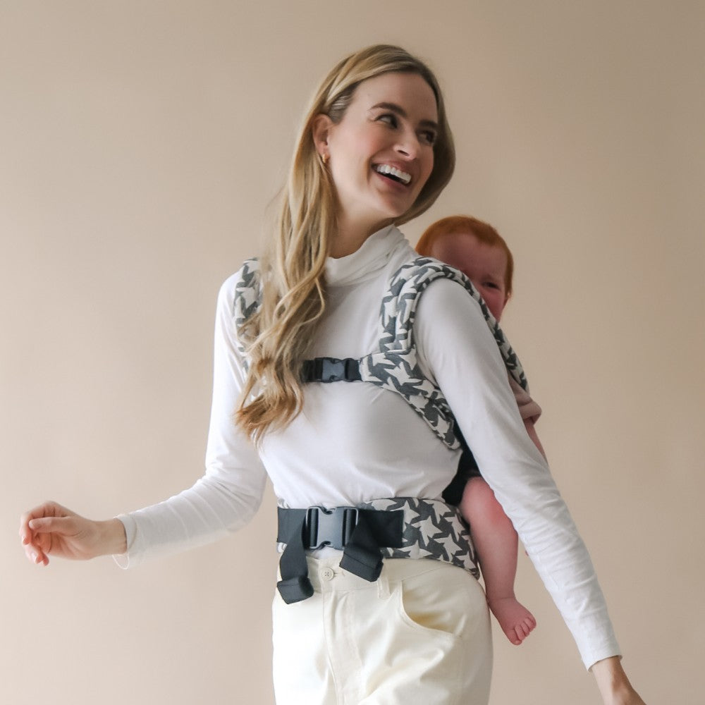 Woman carrying a baby in a patterned baby carrier on her back against a beige background