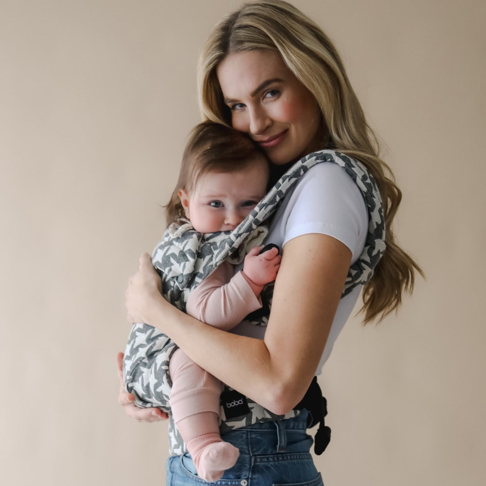On trend high fashion baby carrier worn by a womena in jeans and white tshirt against a beige background