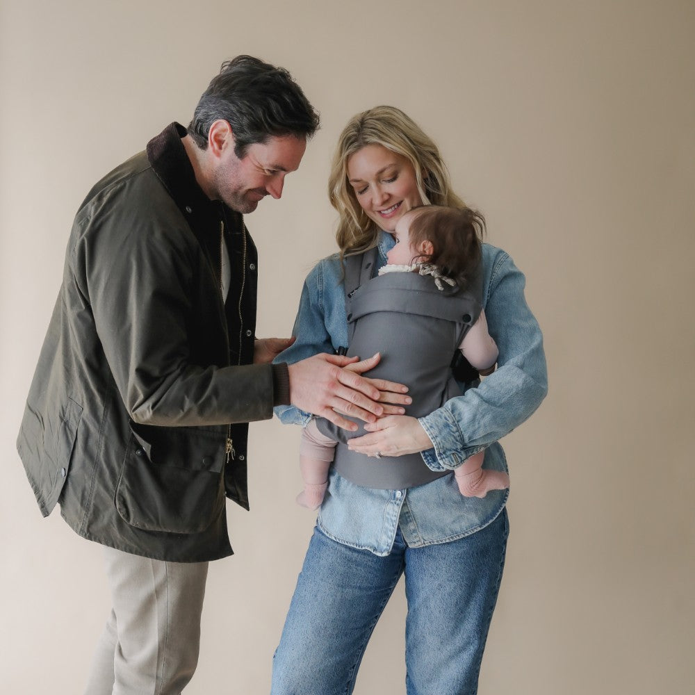 Woman holding a baby in a carrier with a man standing next to them against a beige background