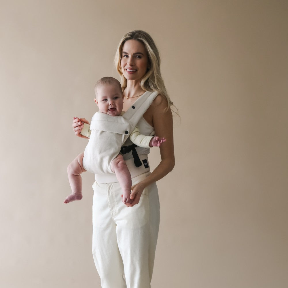 Woman holding a baby in a cream carrier against a beige background in a forward facing position