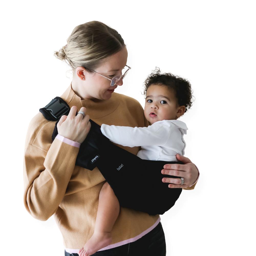 Woman holding a baby in a black side sling baby carrier against a white background