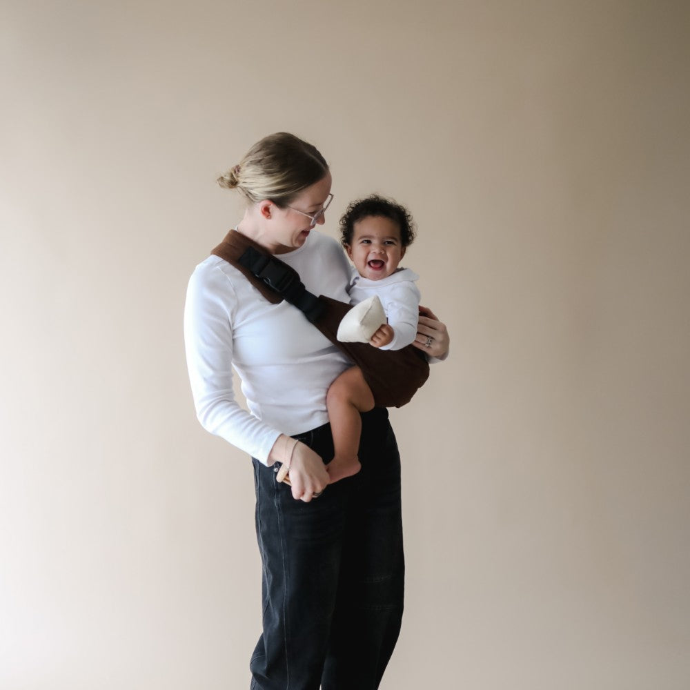Woman holding a toddler in a Boba Buddy sling against a plain background