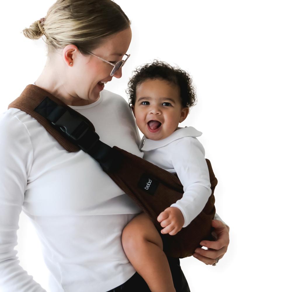 Woman carrying a child in a brown hip side sling carrier against a white background