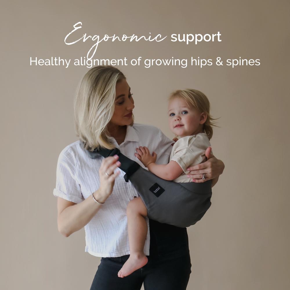 Woman holding a child in an ergonomic toddler hip sling carrier with text about support for growing hips and spines.