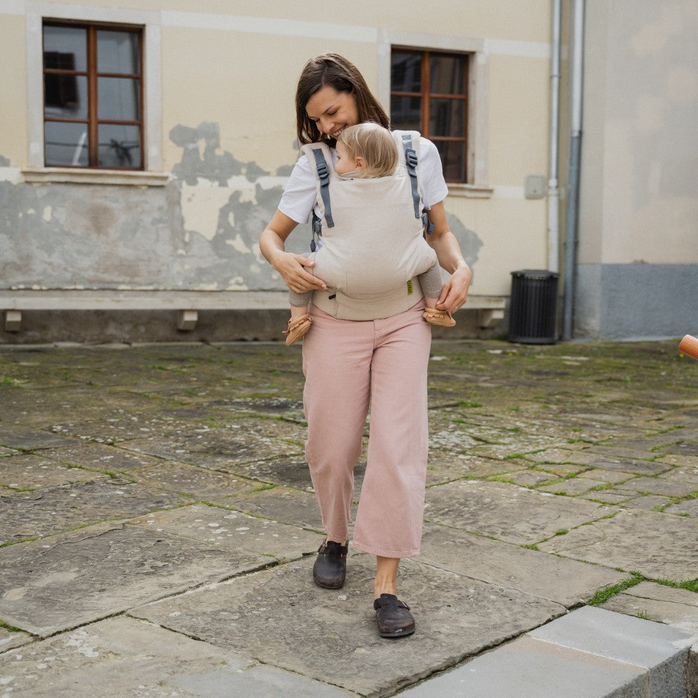 Woman walking outdoors with a child in a Boba X beige linen toddler carrier, wearing a white shirt and pink pants.