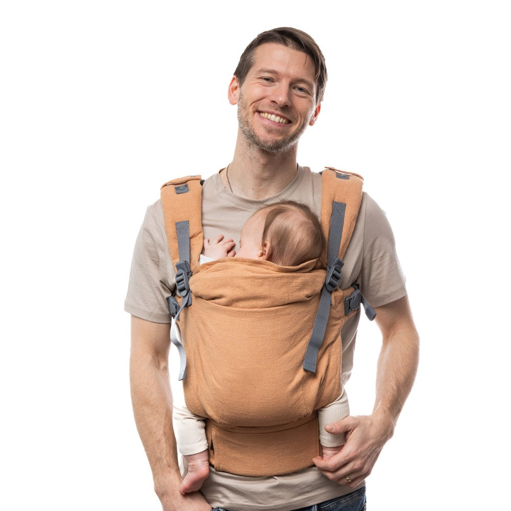 Man carrying a small baby in an rust brown linen Boba X baby carrier on a white background