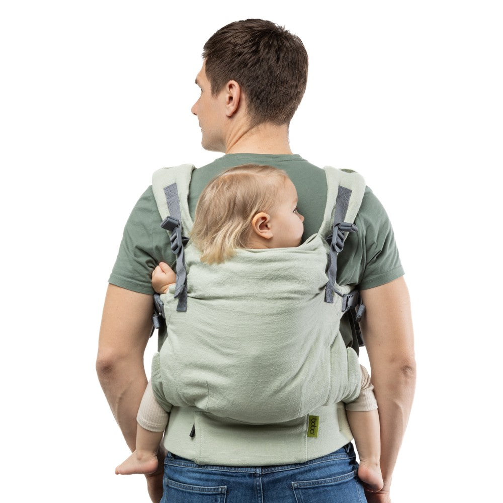 Man carrying a baby on his back in a linen Boba X green baby carrier on a white background