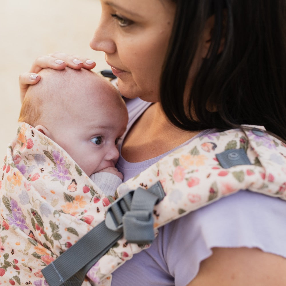 Woman holding a baby in a floral baby carrier showing the adjustable padded straps against a plain background