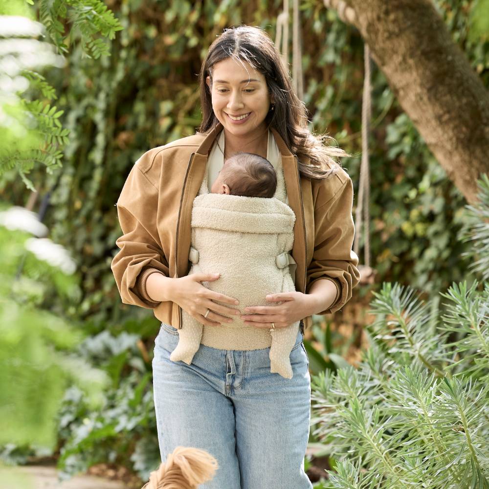 Woman holding a baby in a on trend beige boucle baby carrier outdoors with greenery in the background