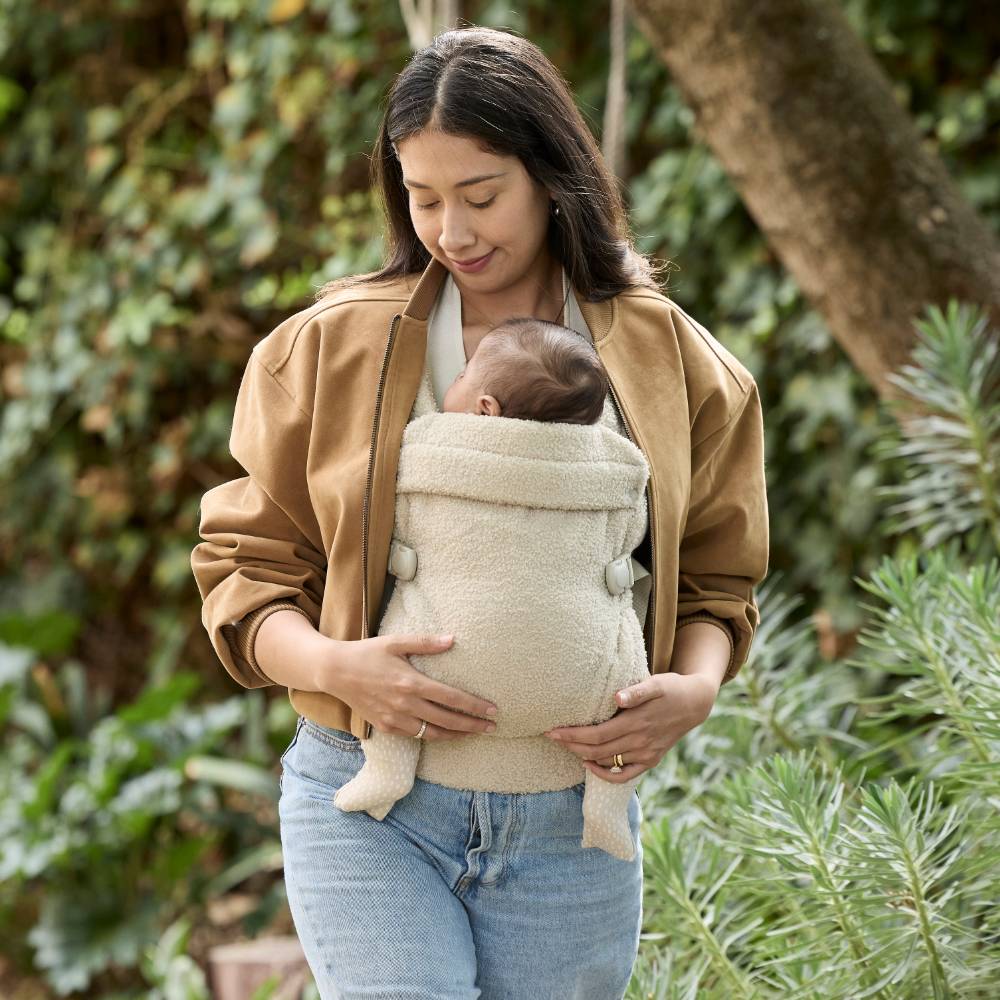 Woman holding a baby in a carrier outdoors with greenery in the background