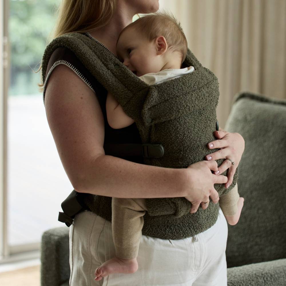 Person holding a baby in a green baby carrier indoors.