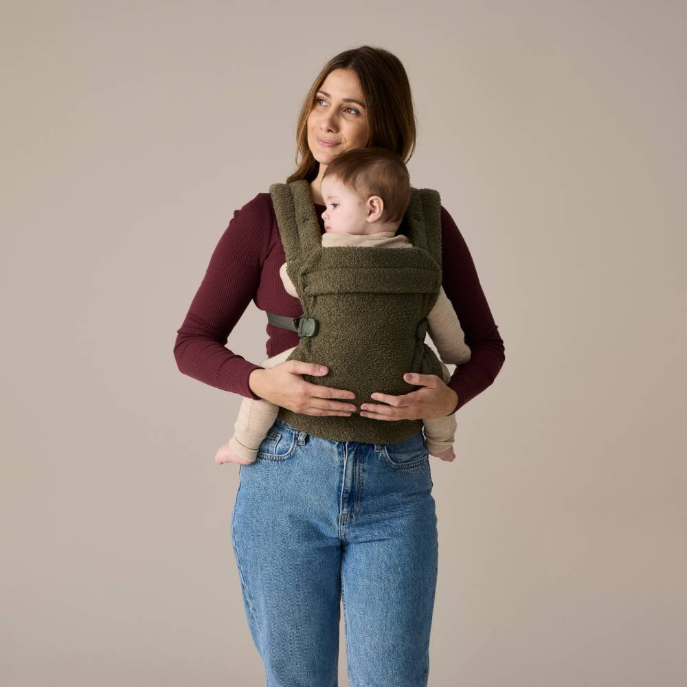 Woman holding a baby in a green baby carrier against a beige background