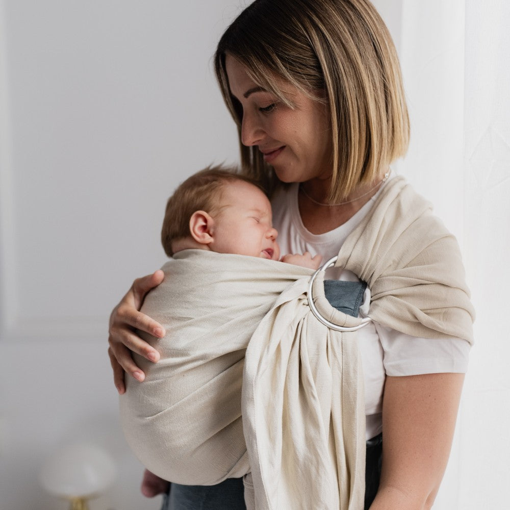 Woman holding a baby wrapped in a linen beige ring sling against a white background