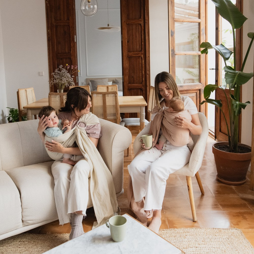 Two women sitting in a living room with babies, holding cups.