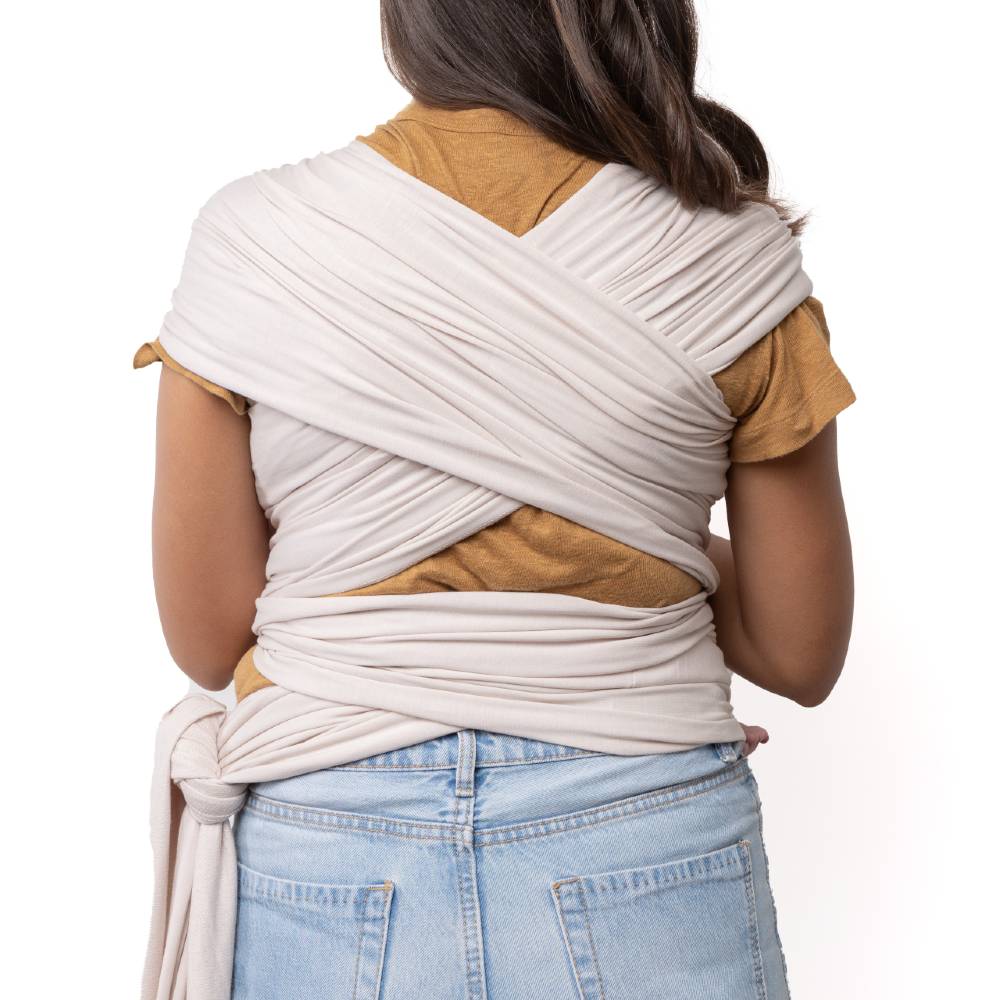 Person wearing a cream organic wrap carrier over a brown top and blue jeans on a white background