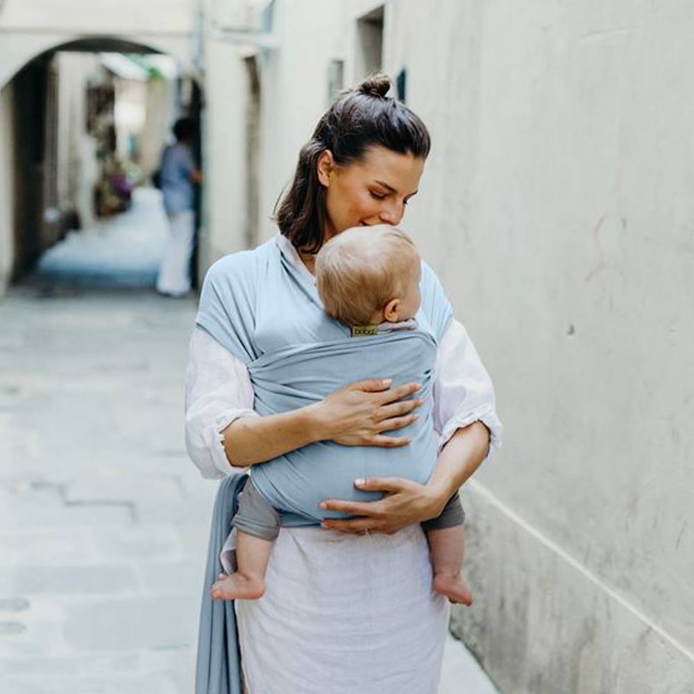 A woman wearing a light blue baby carrier wrap holds and cradles a baby in a real outdoor alleyway setting.