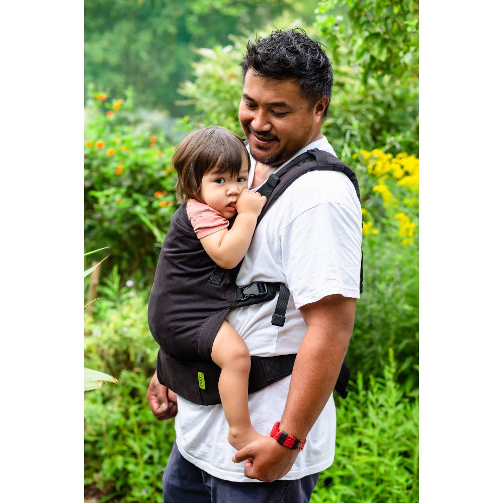 Man holding a child in a black Boba X carrier outdoors with greenery in the background