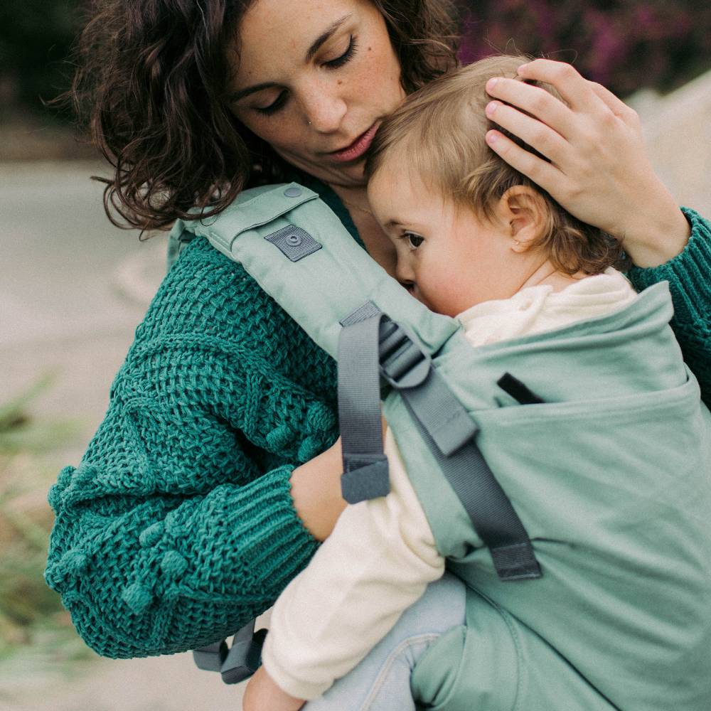 A woman wearing a chunky green knit sweater carries a baby in a sage green front baby carrier. The baby is dressed in a cream long-sleeve top and light blue jeans.
