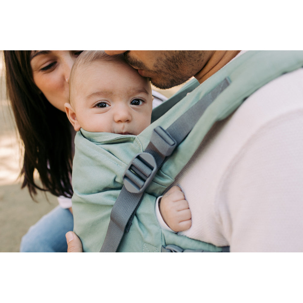 A baby carrier worn on a man's chest with a baby inside, with a woman kissing the man's cheek in the background. The carrier is sage green with gray straps.