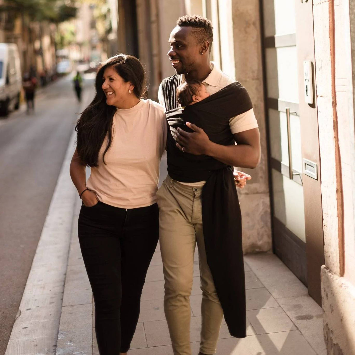 Two people walking down a street, one carrying a child in a sling.