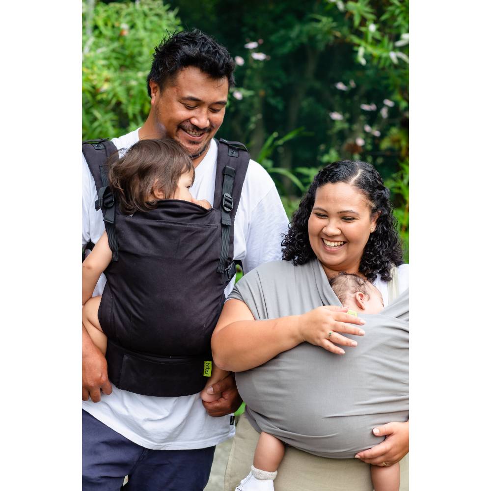 Two parents with their children using baby carriers outdoors.