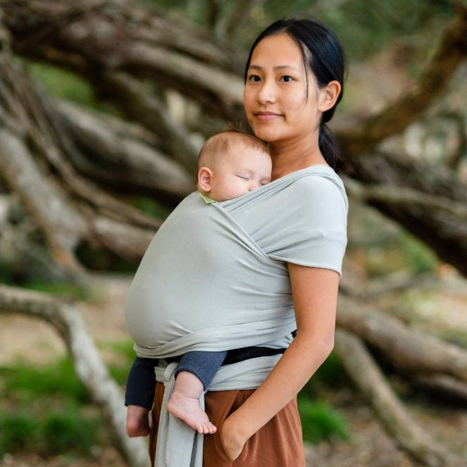 Woman holding a baby in a grey wrap carrier sling outdoors with trees in the background