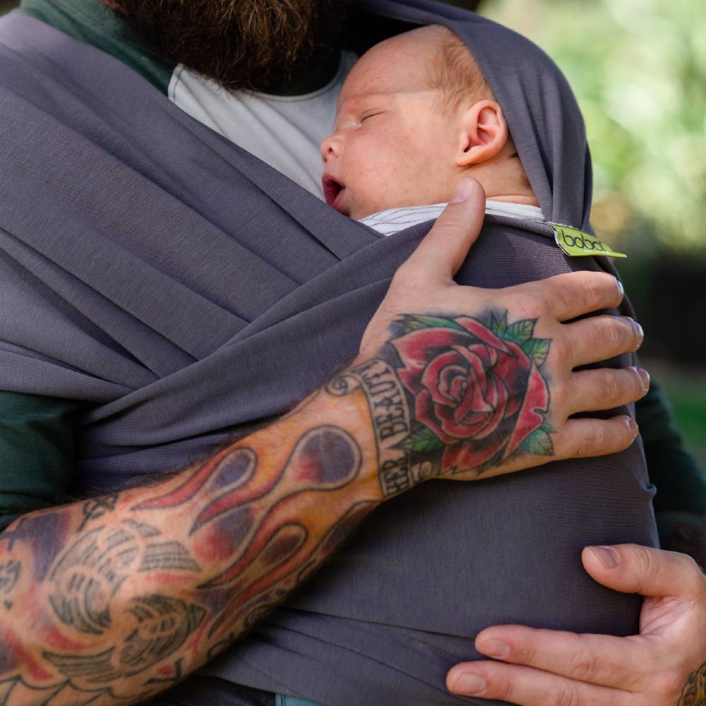 Man holding a newborn baby in a sling with visible tattoos on the arm.