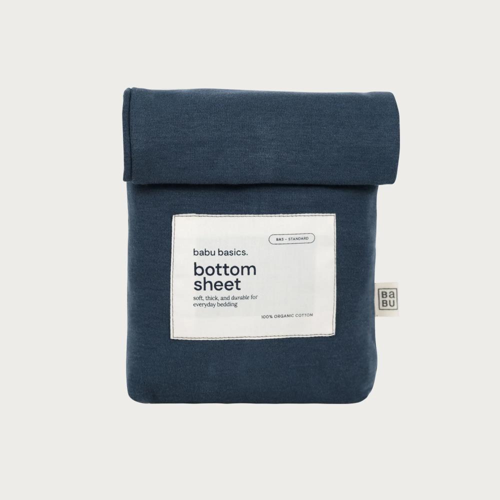 A navy blue bottom sheet by babu basics, neatly folded and rolled at the top, featuring a white paper label on the front with product details and a small brand tag on the right side, set against a light gray background.