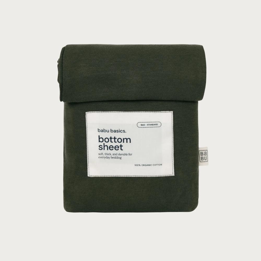 An olive green folded cotton bottom sheet with a white paper label reading 'babu basics. bottom sheet' on a light gray background.
