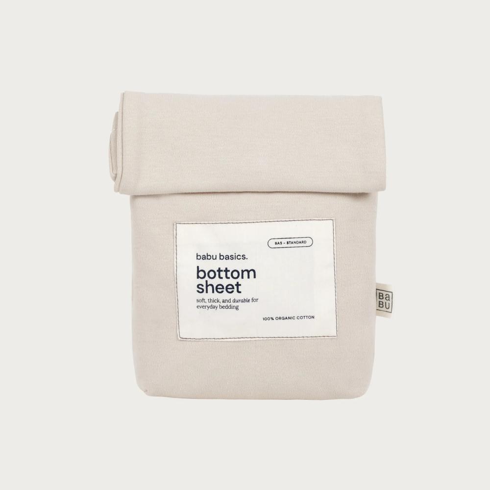 A folded beige cotton bottom sheet with a white paper label reading 'babu basics. bottom sheet' on a light gray background.