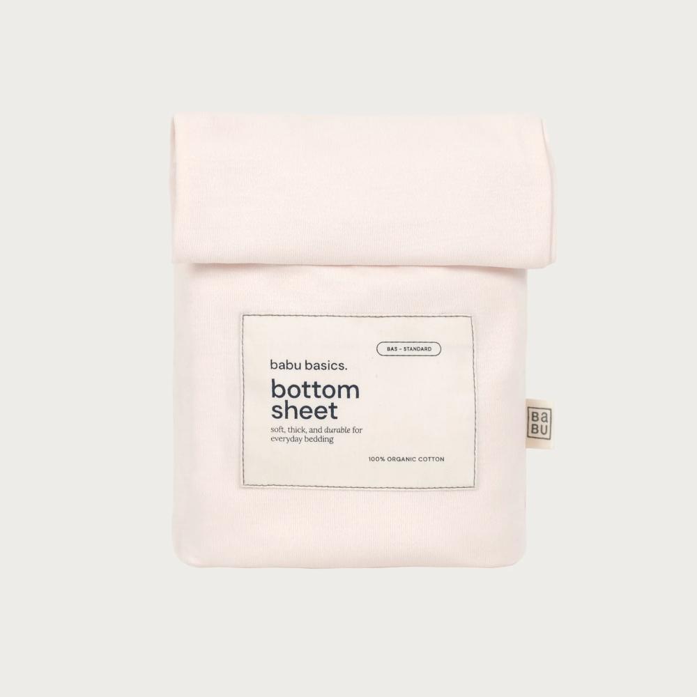 A pale blush pink cotton bottom sheet in a folded pouch package, featuring a white rectangular label with black text that reads 'babu basics. bottom sheet' and '100% organic cotton'.