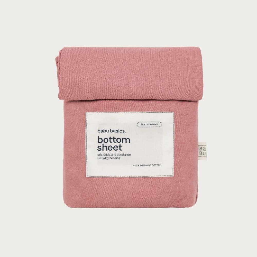 A folded dusty pink cotton bottom sheet by babu basics, neatly rolled and packaged with a white paper label on a light gray background.