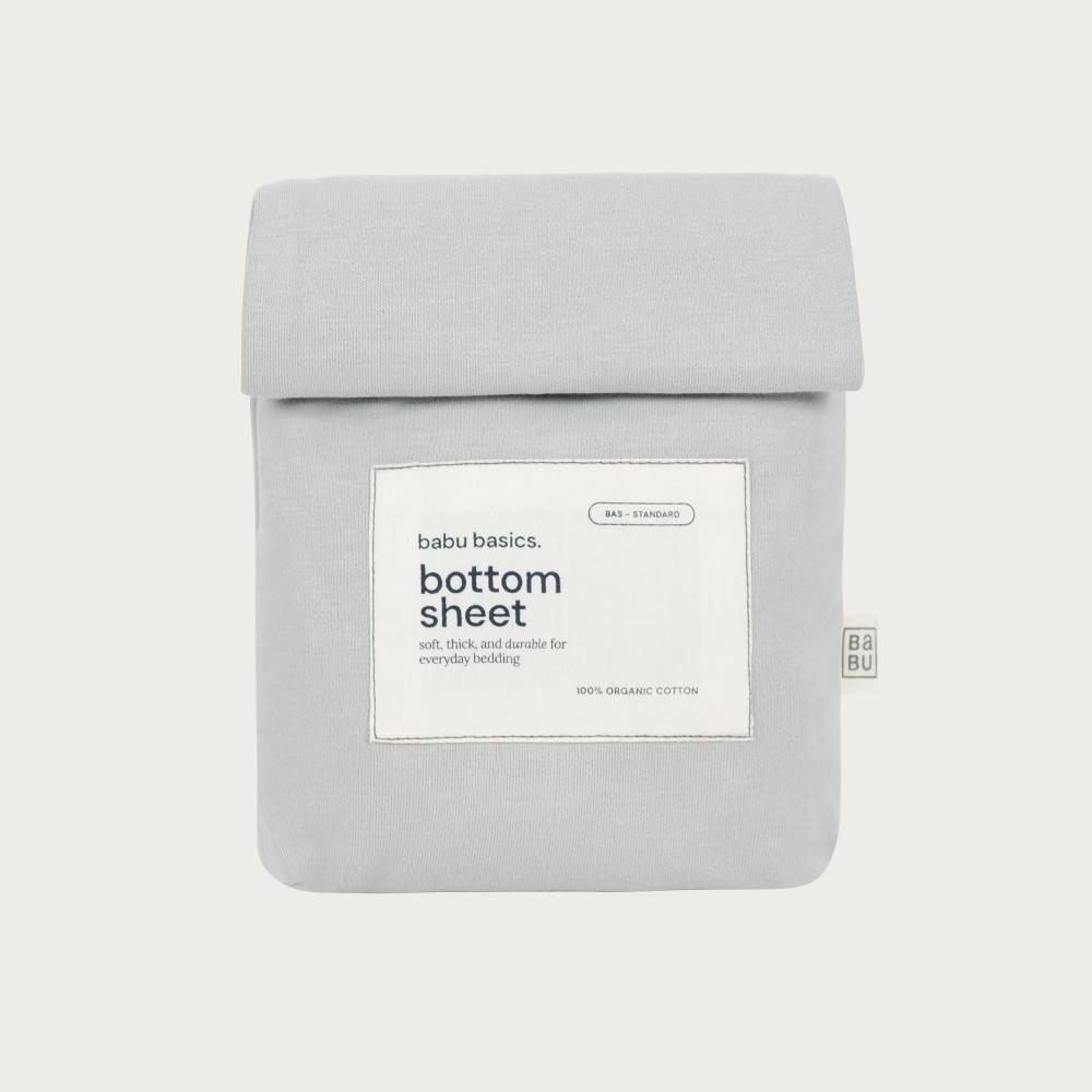 A light gray cotton bottom sheet by babu basics, neatly folded and packaged in a matching light gray sleeve with a white label on the front.