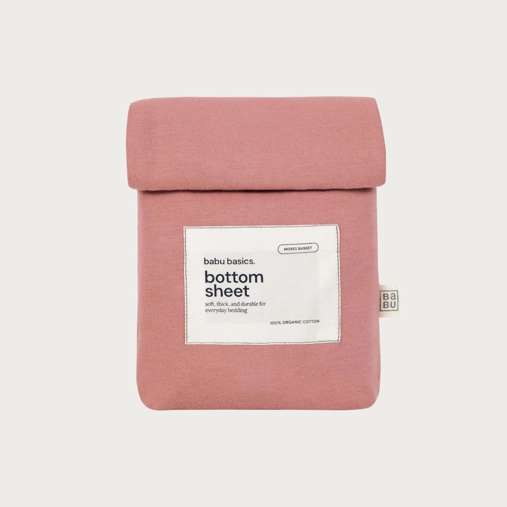 A dusty rose pink folded bottom sheet by Babu Basics, shown against a light gray background. The sheet has a white paper label on the front with black text reading 'babu basics.