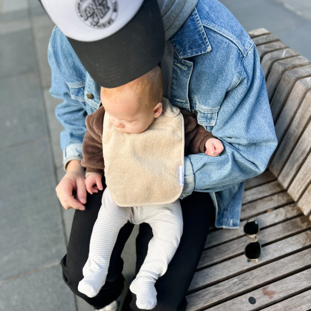 Person holding a baby on a bench outdoors wearing a beige or oat coloured classic style terry towelling bib
