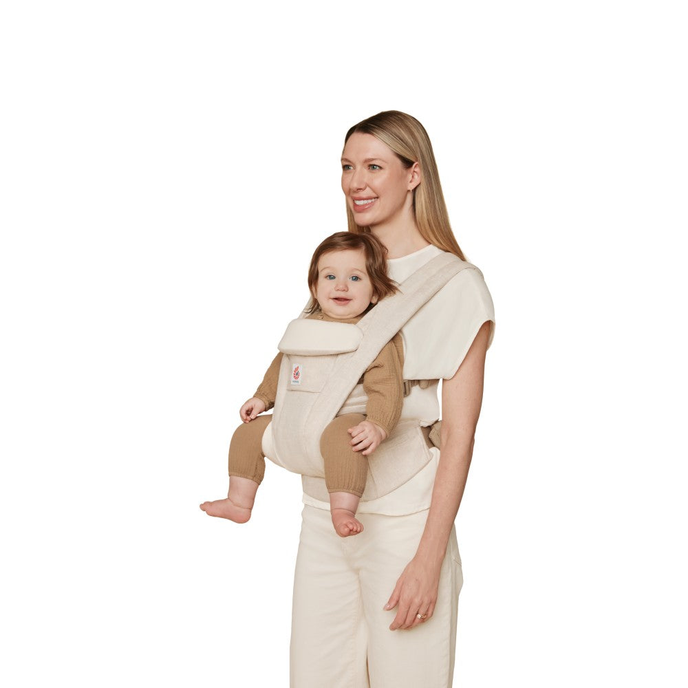 A woman wearing a beige baby carrier with a baby in a tan outfit, photographed against a solid white background.
